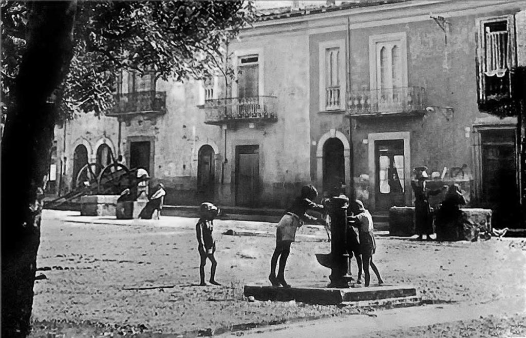 Foto in bianco e nero: donne con conche sulla testa, bambini intorno ai boccali dei pozzi e uomini fermi a riposare